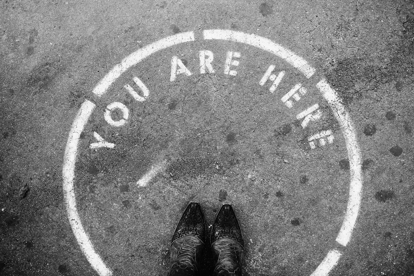 Looking down at a pair of worn boots standing inside a white circle stencilled on tarmac, the words 'YOU ARE HERE' curving around its edge.