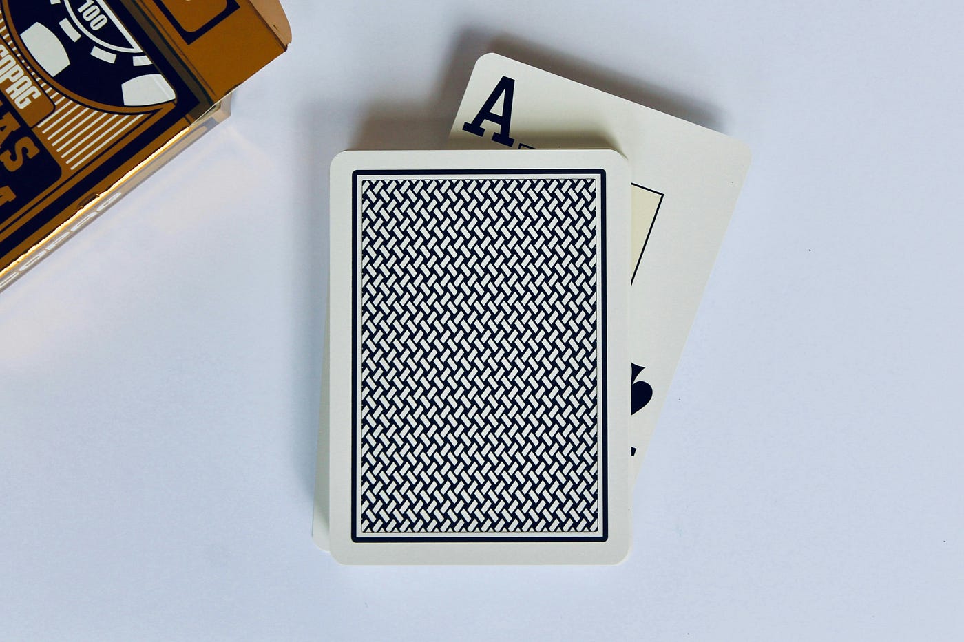 Two playing cards on a white surface beside their box — the top card face down, an ace only half visible beneath it.