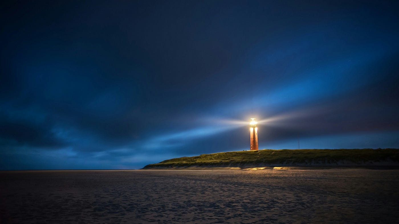 A lighthouse on a low dune at night, its beam cutting a wide shaft of light through deep blue cloud over an empty beach.