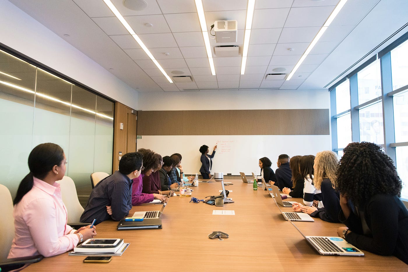 A long boardroom table lined with people and open laptops, one person standing at a whiteboard presenting to the room.