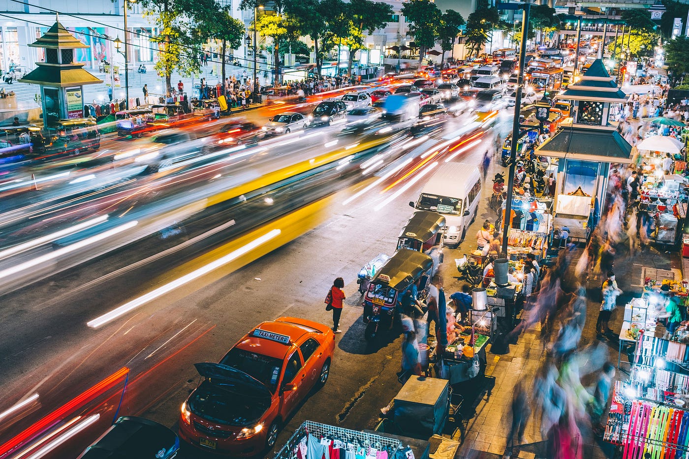 A long-exposure night shot of a crowded city street: traffic reduced to streaks of light, market stalls and pedestrians blurred into motion, with one stationary parked taxi in the foreground.
