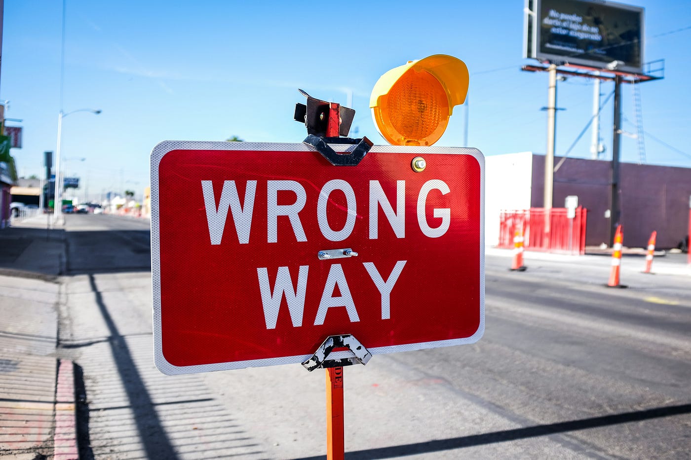 A red 'WRONG WAY' road sign with an amber warning lamp bolted to the top, standing at a sunlit street intersection.
