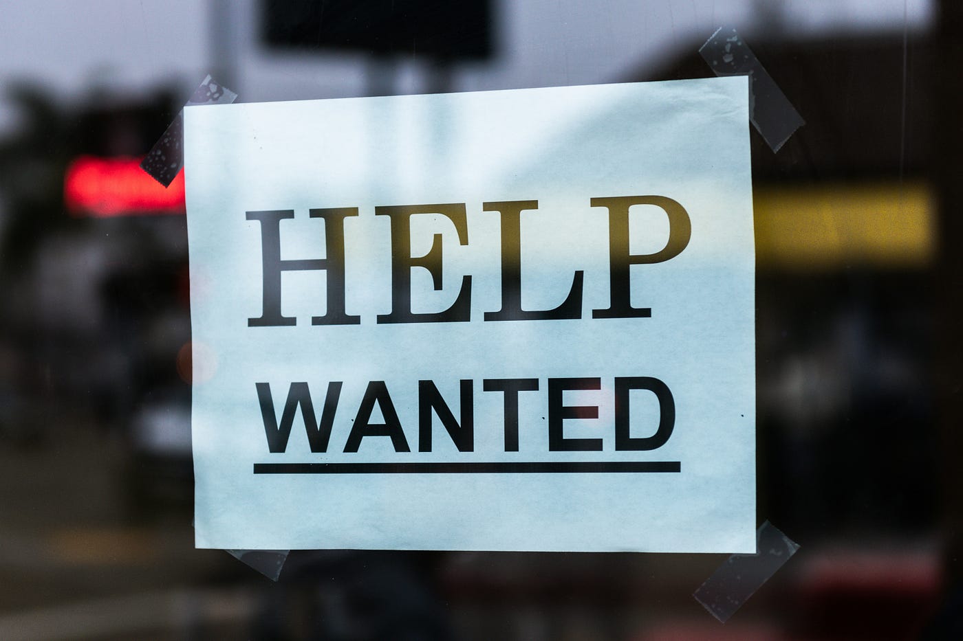 A hand-taped 'HELP WANTED' sign in a shop window, the street behind it out of focus.