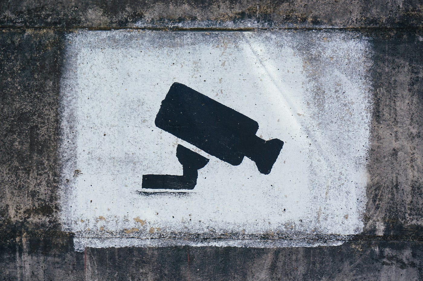 A CCTV camera stencilled in black paint on a whitewashed patch of weathered concrete wall.