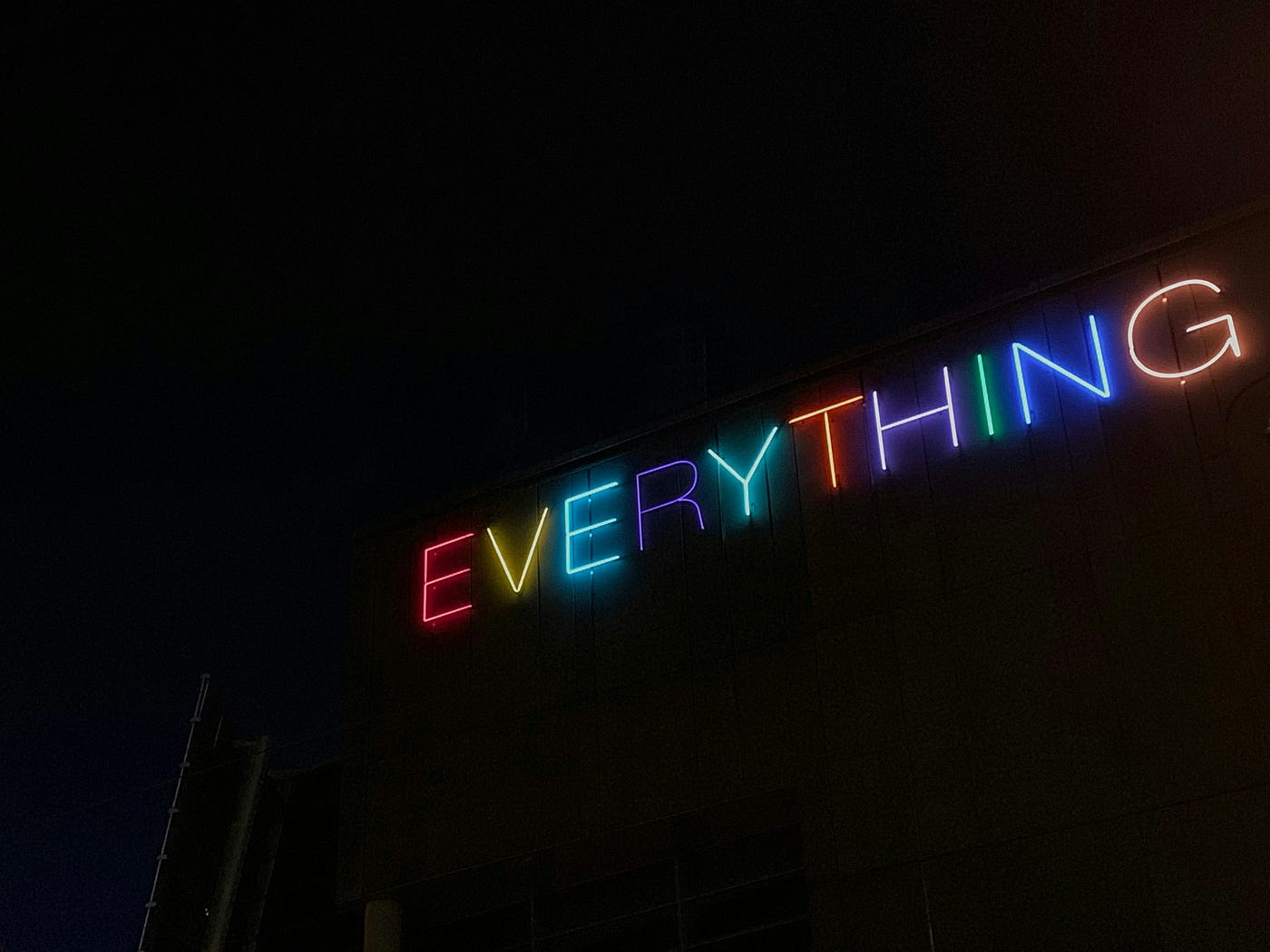 The word 'EVERYTHING' spelled out in multicoloured neon tubing along the roofline of a dark building at night.