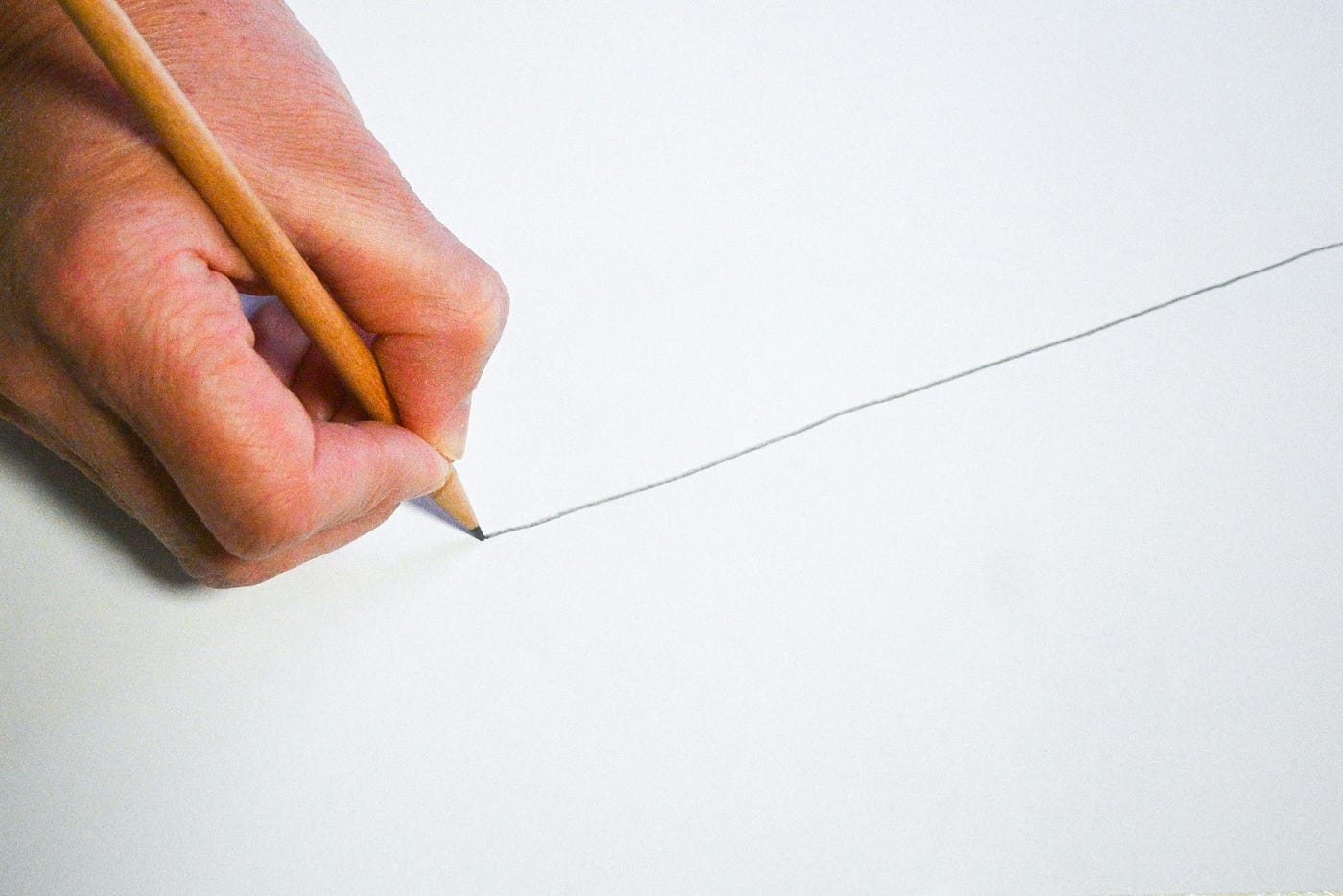 A hand drawing a single unbroken pencil line across a sheet of blank white paper.