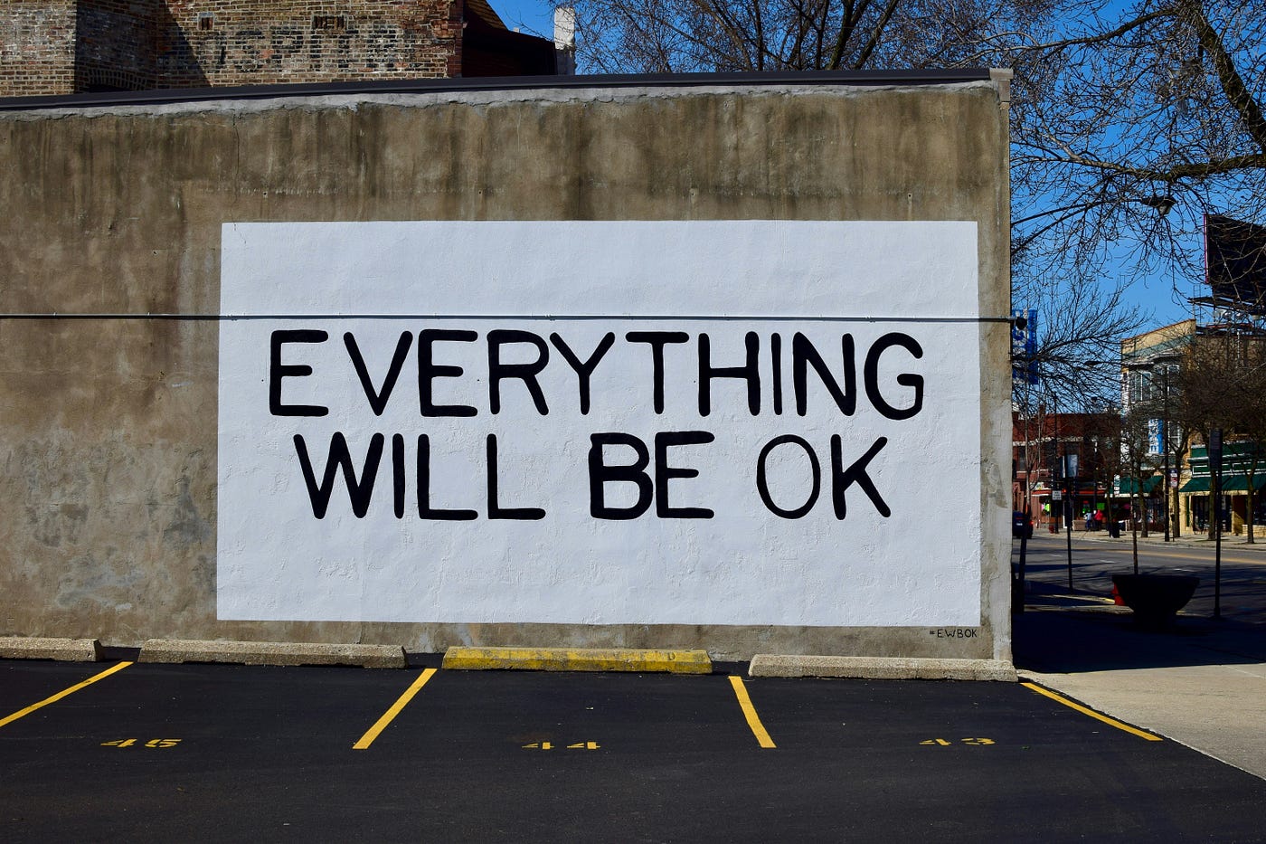 A large white panel painted on the side of a building beside an empty car park, hand-lettered in black capitals: 'EVERYTHING WILL BE OK'.