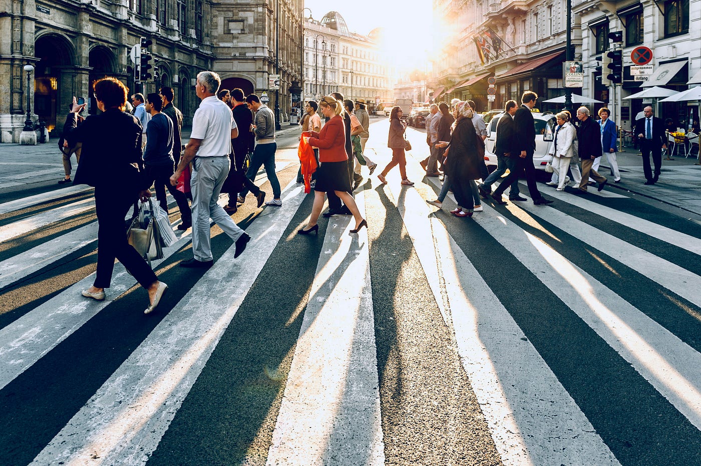 A crowd of pedestrians crossing a wide city street on a zebra crossing at sunset, long shadows stretching toward the camera.