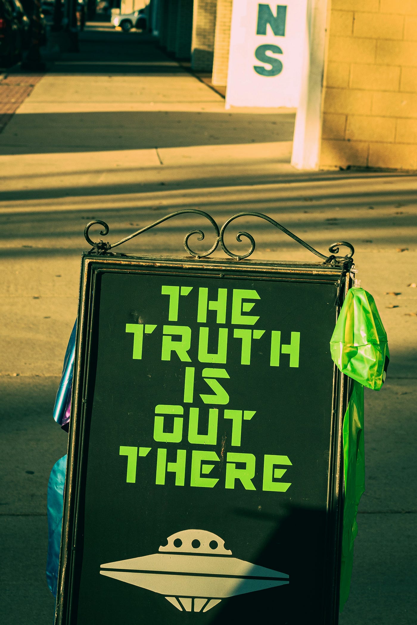 A pavement A-frame sign on a sunlit street reading 'THE TRUTH IS OUT THERE' in green stencil letters above a flying saucer.