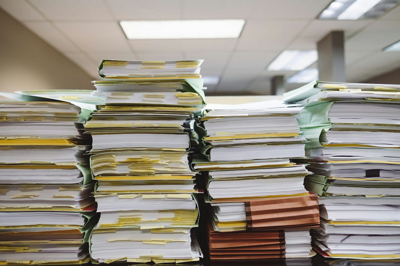 Towering stacks of paper files and folders bristling with yellow index tabs, piled on a desk in a strip-lit office.