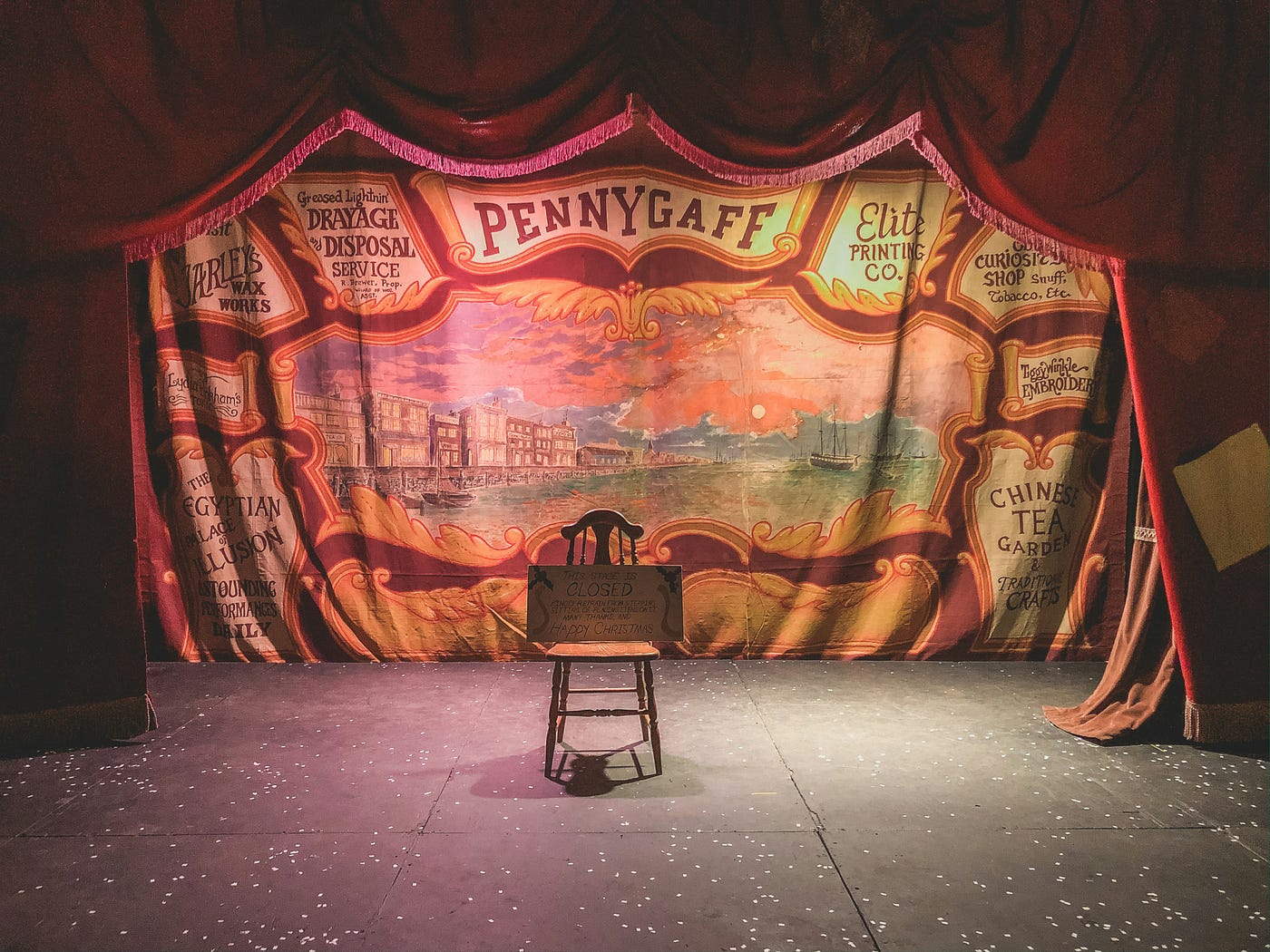 An empty Victorian music-hall stage with a painted advertising backdrop, a single wooden chair centre stage holding a hand-lettered sign reading 'This stage is closed'.
