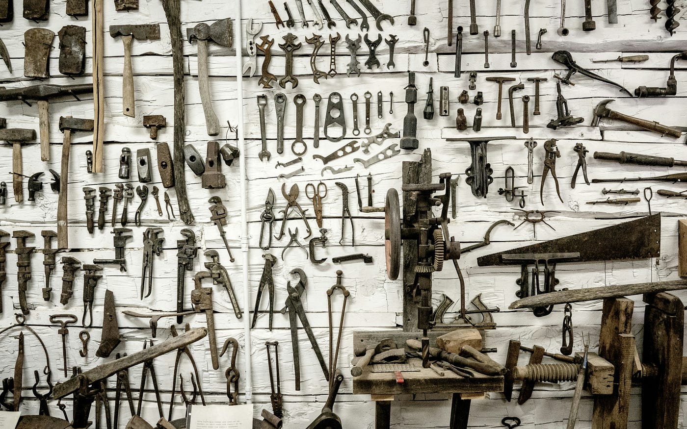 A whitewashed workshop wall covered edge to edge with dozens of old hand tools — wrenches, hammers, saws and pliers — hung in rows.