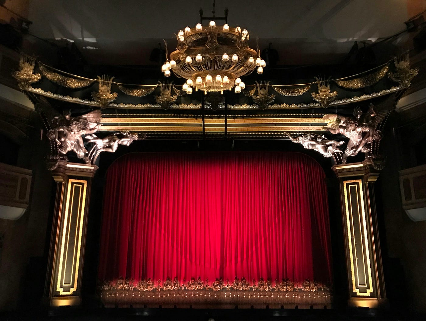 An ornate gilded theatre proscenium lit by a chandelier, with the red velvet curtain still closed across the stage.