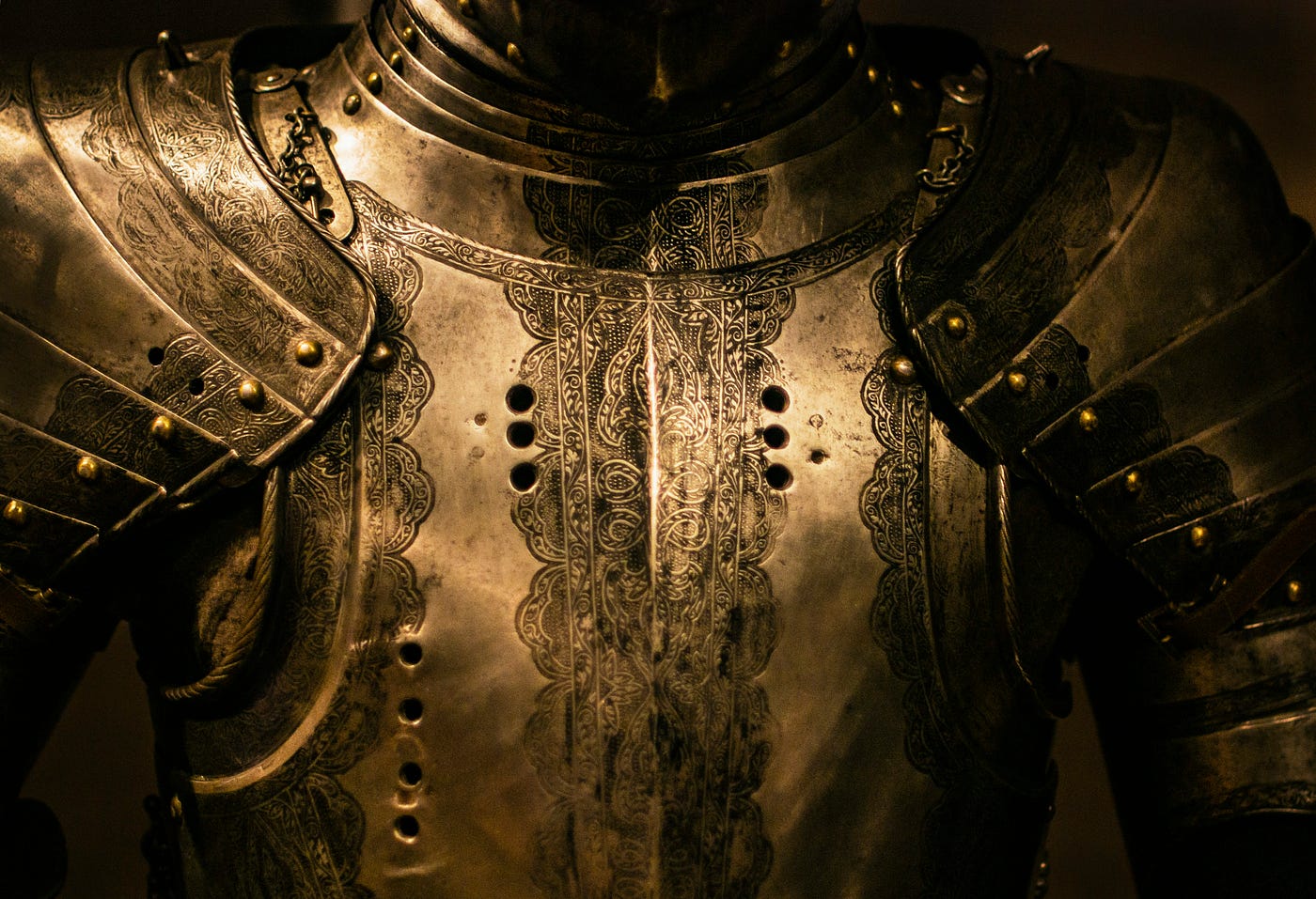 The engraved breastplate and shoulder plates of a suit of antique armour, lit warmly against a dark background.